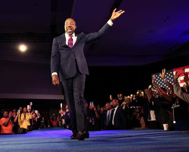 A black man dressed in a dark business suit is walking on a stage and waving to hundres of people clapping their hands.