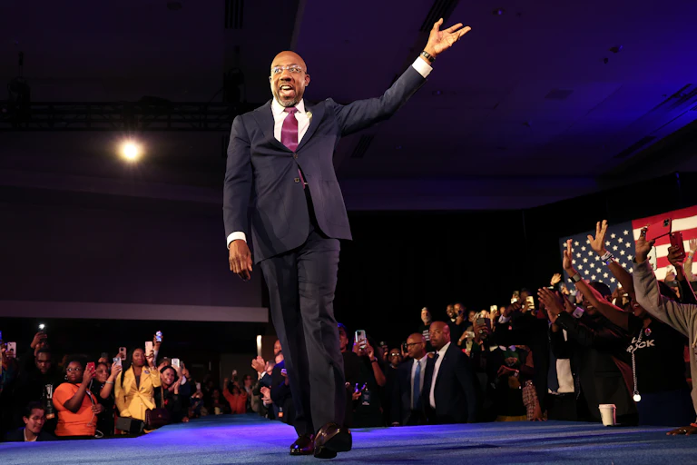 A black man dressed in a dark business suit is walking on a stage and waving to hundres of people clapping their hands.