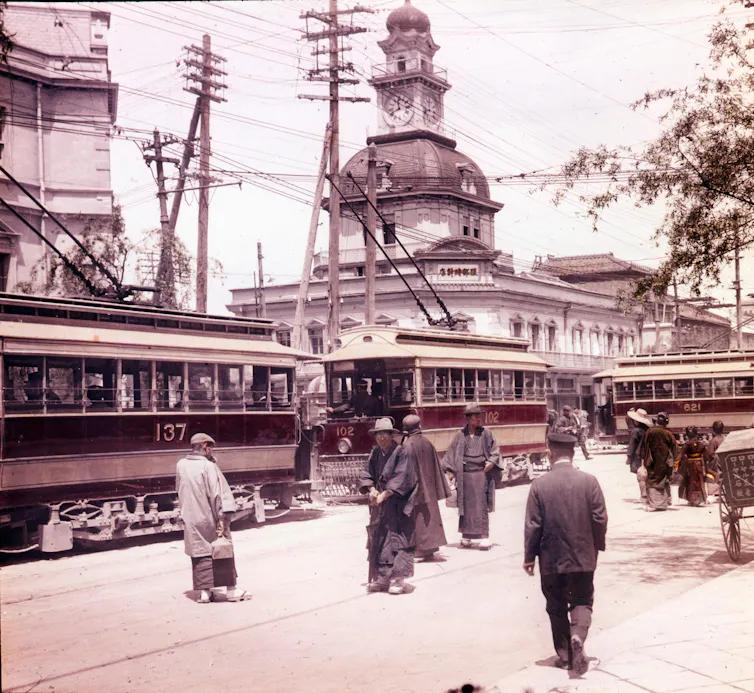 Colorized photo of pedestrians and streetcars with a clock tower in the background.