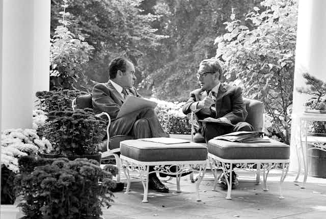 Two men in suits sit on a porch and talk.