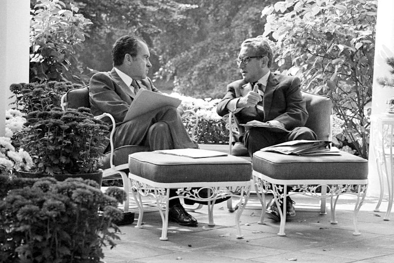 Two men in suits sit on a porch and talk.