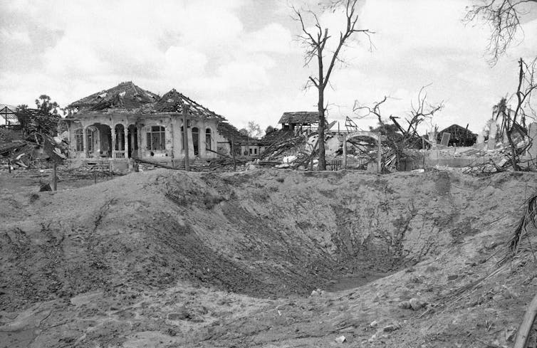 A large crater in the ground with burned trees and ruined buildings behind it.