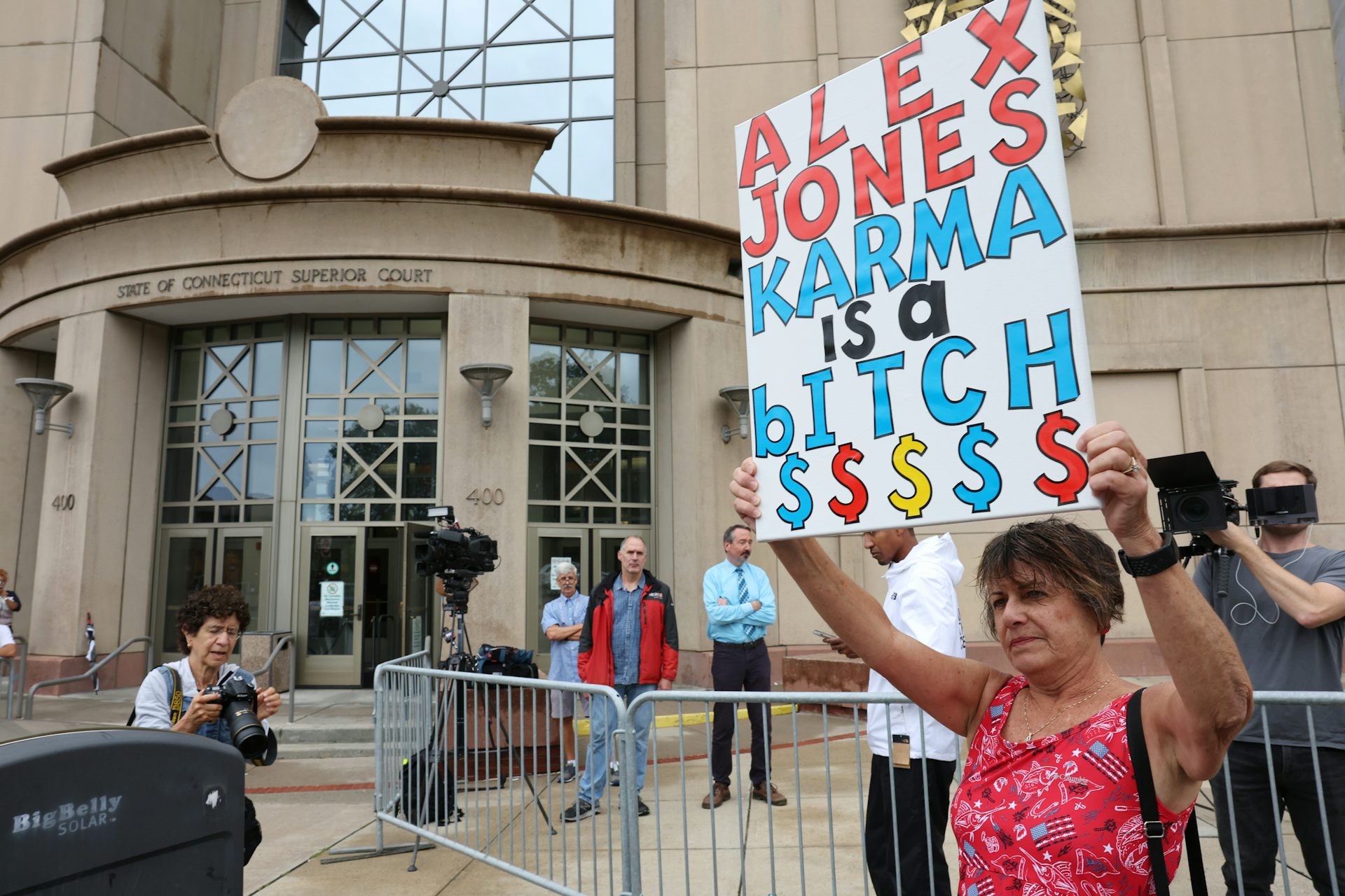 Woman stands outside courthouse in Waterbury, Connecticut holding sign above her head that reads, 'Alex Jones, Karma is a bitch,' with dollar signs at the bottom.