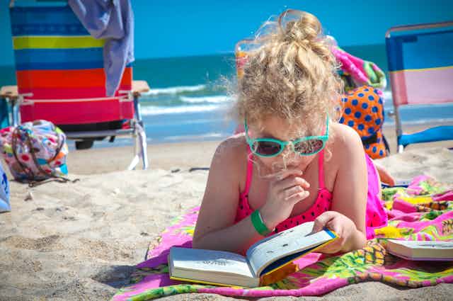 primary school aged girl reading on a beach towel on sand, wearing sunglasses