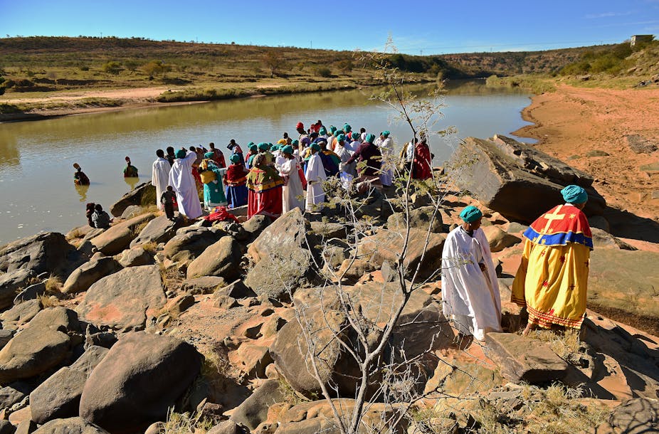 People wearing robes stand on a river bank. Others are in the water.