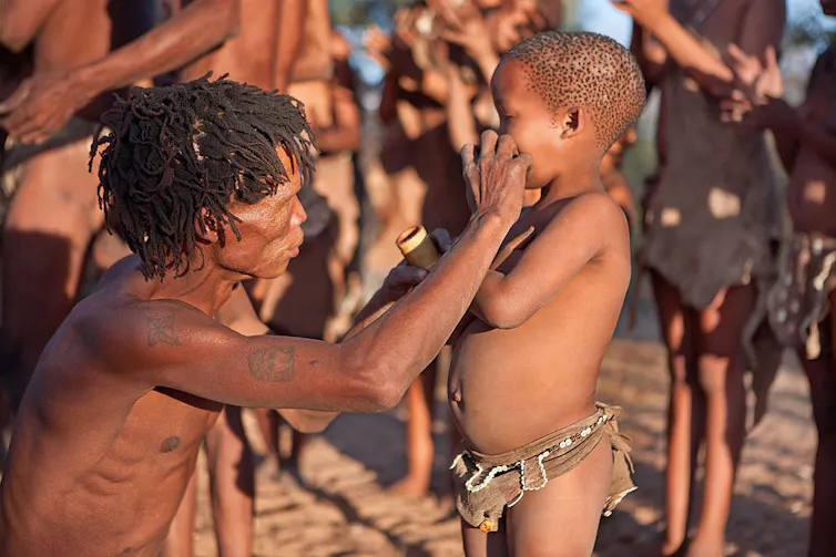 A man in traditional hunting clothing crouches to apply paint with his finger on a boy child's cheeks.