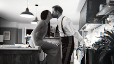 Black and white photograph of Meghan sat on a kitchen counter in a gown and Harry in a suit without his jacket leaning in to kiss her.