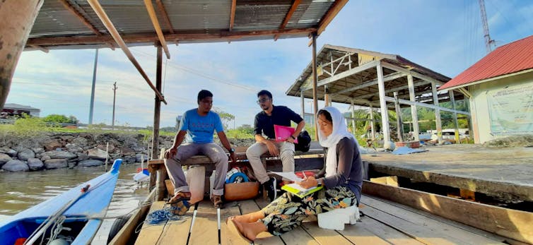 Três pessoas sentadas à beira d'água conversando entre si.