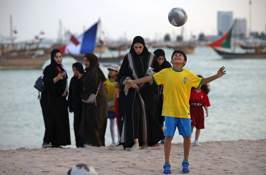Garçon jouant au football sur la plage. Au second plan, plusieurs femmes voilées.