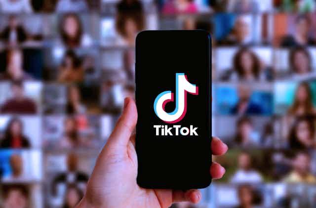 A hand holds a phone with Tik Tok loaded up in front of a blurred background of faces on other screens.