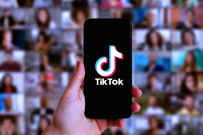 A hand holds a phone with Tik Tok loaded up in front of a blurred background of faces on other screens.