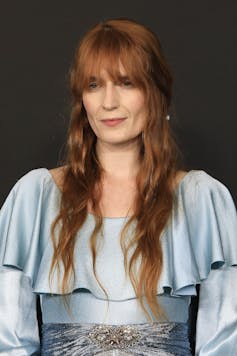 Portrait photo of Florence Welch. She has long red hair down to her waist with a full fringe and wears a light blue dress with a frilled cape top.
