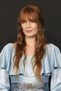 Portrait photo of Florence Welch. She has long red hair down to her waist with a full fringe and wears a light blue dress with a frilled cape top.