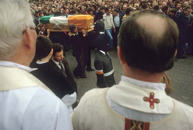 Two Catholic clergy watch as a coffin goes by draped in the Irish tricolour.