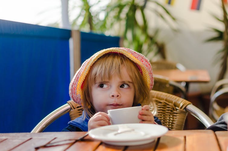 Une petite fille coiffée d’une capeline se penche sur une table et tient une tasse à café d’un air malicieux