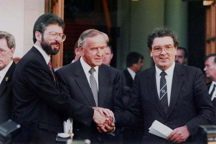 Three men in black suits - Gerry Adams, Albert Reynolds and John Hume - shaking hands.