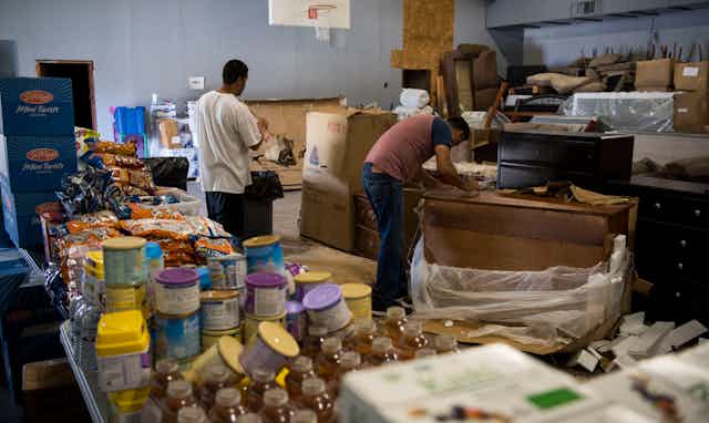 Two men sort through donated food and furniture in a warehouse.