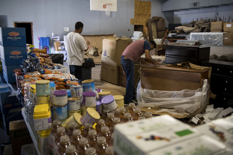 Two men sort through donated food and furniture in a warehouse.