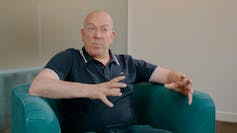 Terry Stone wears a navy blue polo shirt and sits on a green couch.