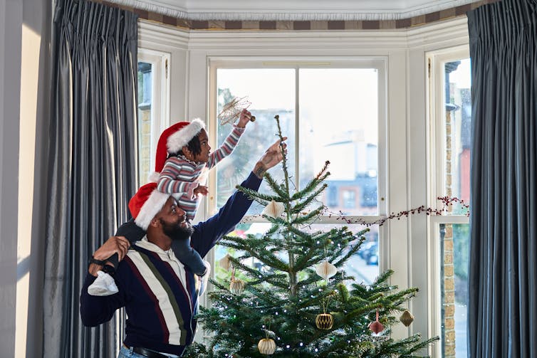 child on mans shoulders putting star atop Christmas tree