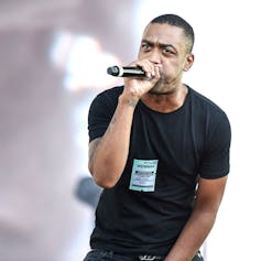 Rapper Wiley performing on stage in a black tshirt, rapping into a microphone.