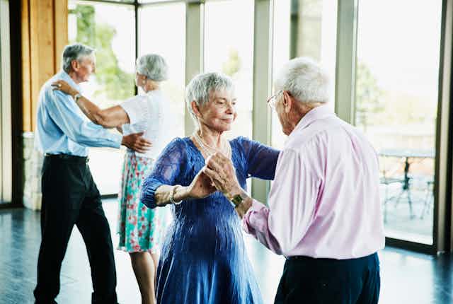 Two well-dressed elderly couples engage in ballroom dancing.