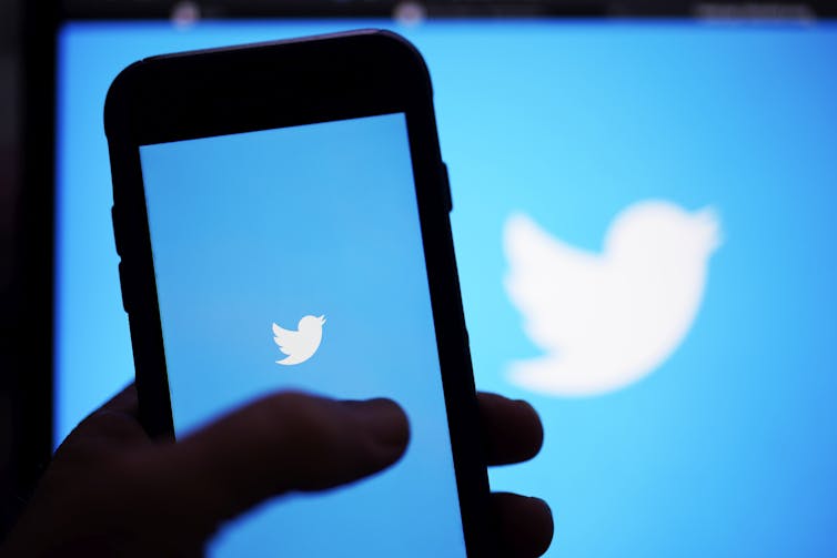 A hand holds a phone with the Twitter logo on it in front of a screen that also has the Twitter logo on it