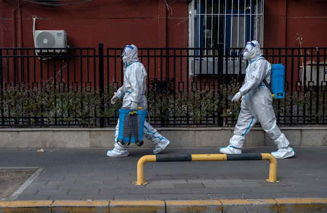 Two workers in protective suits carrying disinfection equipment.