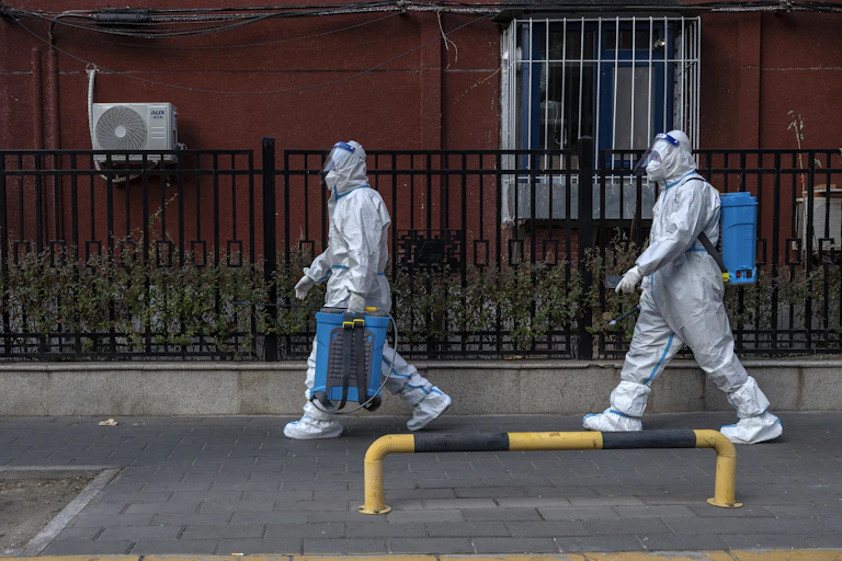 Two workers in protective suits carrying disinfection equipment.