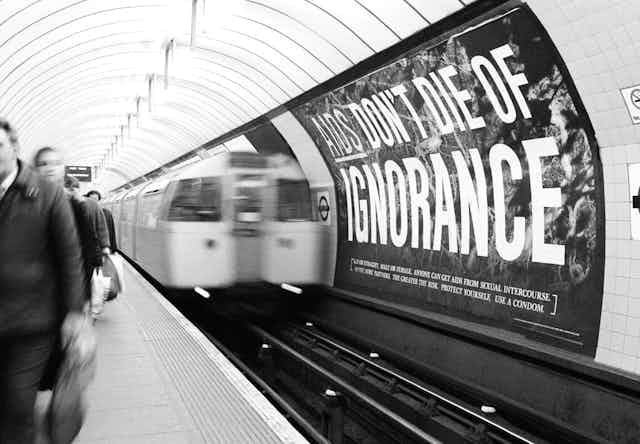 Subway riders passing poster that reads "AIDS don't die of ignorance"