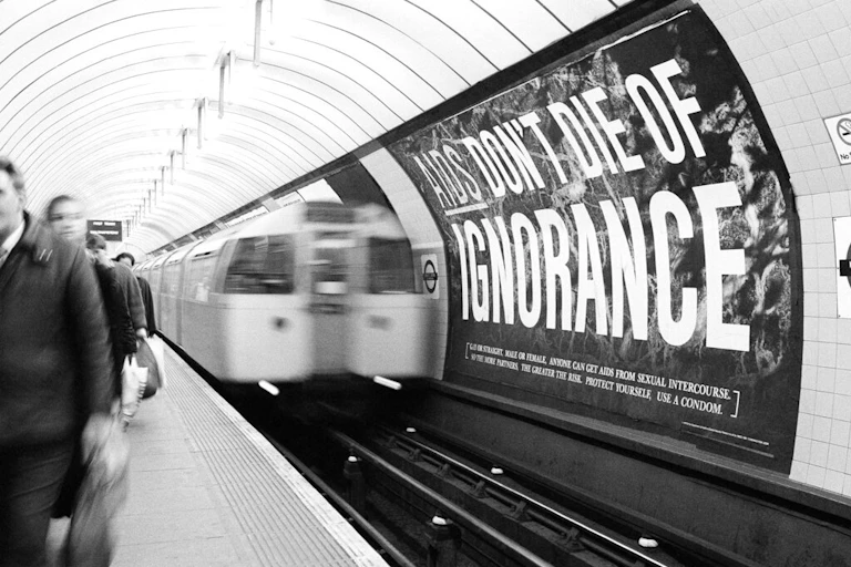 Subway riders passing poster that reads "AIDS don't die of ignorance"