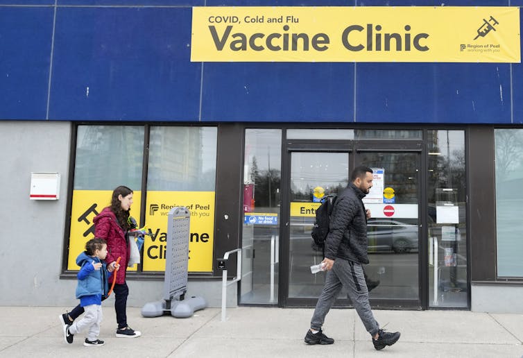 People walk past a blue storefront with a yellow Vaccine Clinic sign
