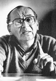 A black and white portrait of a man with a sweater, glasses and thinning fair, holding up his finger.