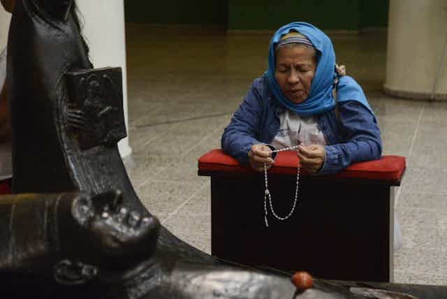A woman with a blue scarf on her head kneels while praying the rosary in front of a statue.
