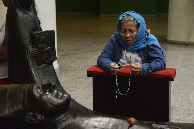 A woman with a blue scarf on her head kneels while praying the rosary in front of a statue.