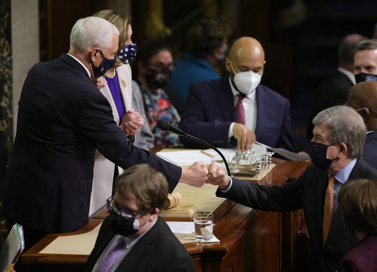 A man in a dark suit with white hair bumps fists with another man in a dark suit with gray hair.