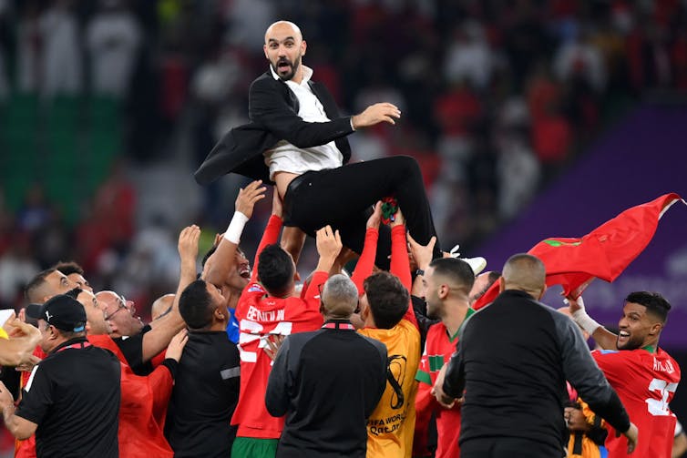 Seorang lelaki berjas dan berkemeja putih diangkat tinggi ke udara oleh sekelompok laki-laki yang mengenakan pakaian sepak bola berwarna merah dan hijau Maroko.