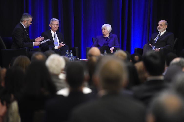 Two white men and a white woman sit in chairs on a stage in front of an audience as another man stands nearby