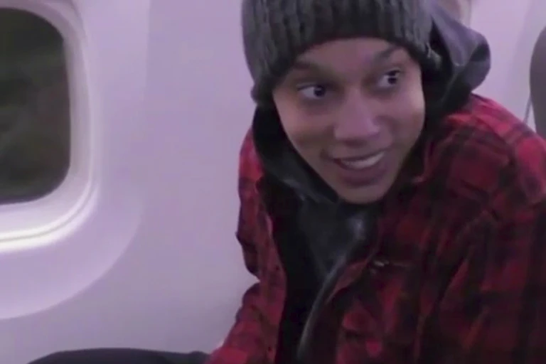 A woman in a winter hat and a checked jacket smiles while sat in an airplane seat.