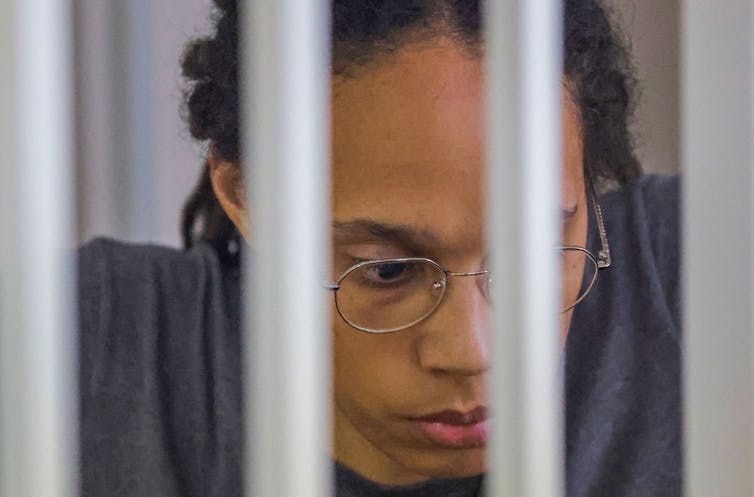 A woman in glasses looks pensive behind bars.