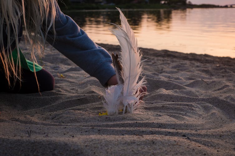 A young child makes a mandala in the sand with feathers.