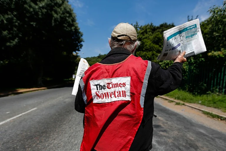 A man wearing a high-visibility red jacket written The Times Sowetan on the back holding up a newspaper at a street intersection.