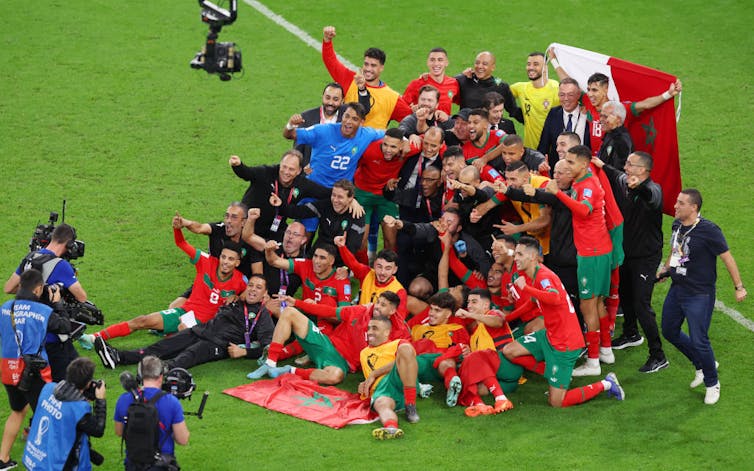 A football team poses in the middle of a field with a bank of cameras taping and photographing them.