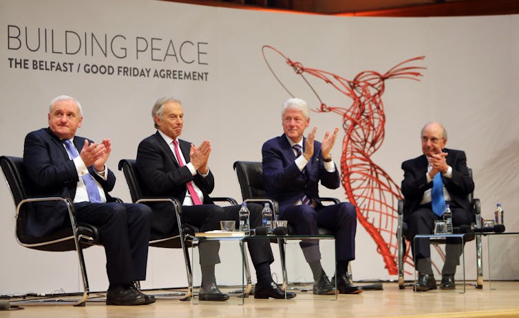 Four men in suits, on a stage, clapping, white background with red sculpture image and writing