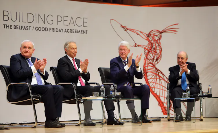 Four men in suits, on a stage, clapping, white background with red sculpture image and writing