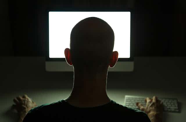 The back of a man's head as he looks at a computer monitor in a dark room.