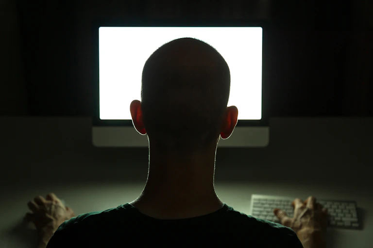 The back of a man's head as he looks at a computer monitor in a dark room.