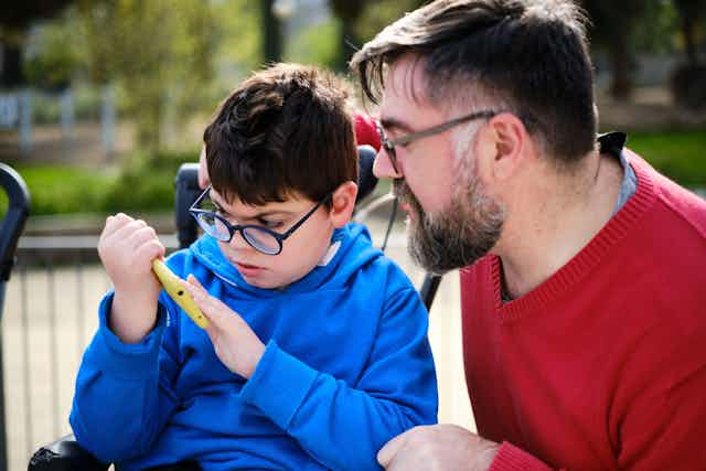 A man in a red jumper and glasses kneels beside a child in a blue jumper in a wheelchair, holding a yellow phone.