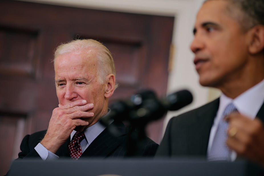 A man wearing a suit and a tie covers his mouth as another to his left, also wearing a suit anda tie, speaks into two microphoes.