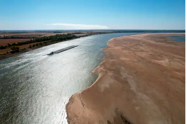 A barge moves through a narrow channel next to a large exposed sandbank